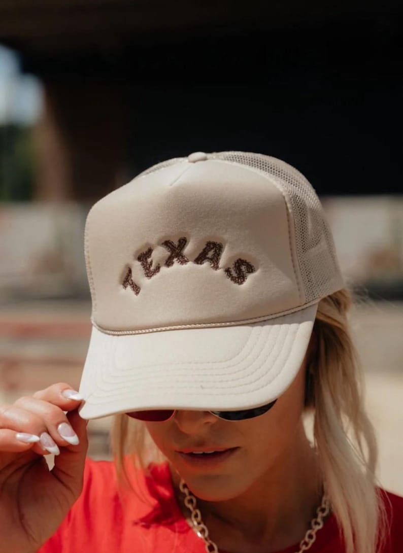 May include: A beige trucker hat with a brown embroidered "TEXAS" logo. The hat has a mesh back and a curved brim.