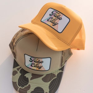 May include: Two trucker hats with a camouflage pattern and a yellow mesh back. Both hats have a patch that says "Take it Easy" with a rainbow design.