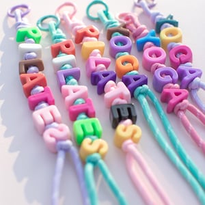 May include: Colorful beaded keychains spelling out names. Each keychain features a different color scheme, with letters in various hues like pink, green, and orange. The keychains are strung on matching cords, creating a playful and personalized accessory.