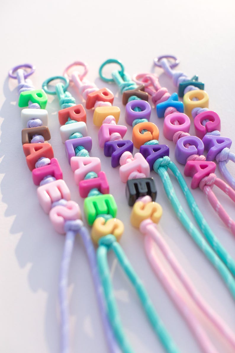 May include: Colorful beaded keychains spelling out names. Each keychain features a different color scheme, with letters in various hues like pink, green, and orange. The keychains are strung on matching cords, creating a playful and personalized accessory.