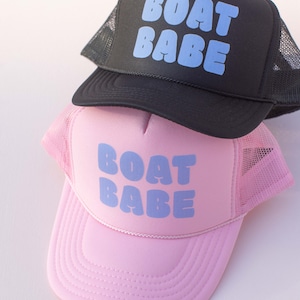 May include: Two trucker hats with the text "BOAT BABE" in blue lettering. The top hat is black with a black mesh back. The bottom hat is pink with a pink mesh back.