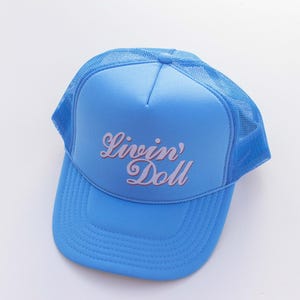 May include: A light blue trucker hat with a mesh back and a curved brim. The hat features the text "Livin' Doll" in a cursive font across the front. The hat is set against a white background.