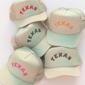 May include: Five beige trucker hats with mesh back panels. Each hat has the word "TEXAS" embroidered on the front in a different colour: red, orange, green, brown, and pink.