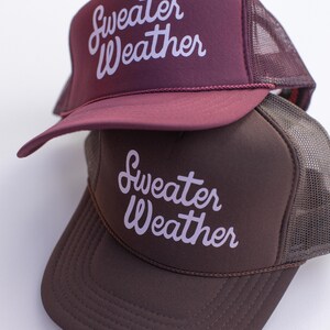 May include: Two trucker hats, one burgundy and one brown, stacked on top of each other. Each hat has a mesh back and a curved brim. The phrase "Sweater Weather" is printed in white script across the front of each hat.