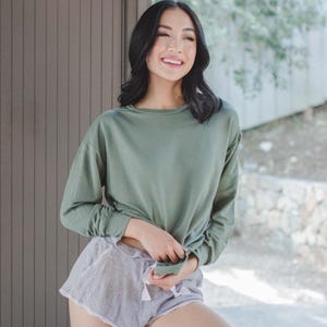 May include: An olive green long-sleeve shirt and striped shorts with a drawstring waist. The shorts have a ruffled hem. The outfit is casual and comfortable, suitable for lounging or light activity.