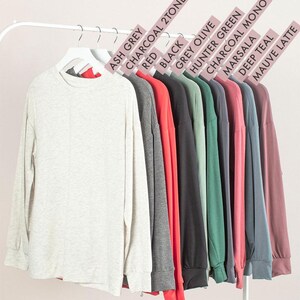 May include: A display of long-sleeved shirts in various colors, including Ash Grey, Red, Black, and Mauve Latte, hanging on a white rack. Each shirt has a crew neck and is made of a soft material. Color labels are visible above each shirt.