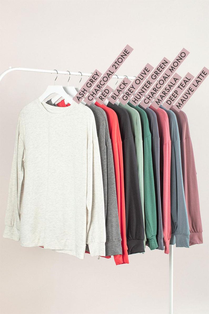 May include: A display of long-sleeved shirts in various colors, including Ash Grey, Red, Black, and Mauve Latte, hanging on a white rack. Each shirt has a crew neck and is made of a soft material. Color labels are visible above each shirt.