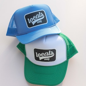 May include: Two trucker hats with a black and white "locals only" logo. The top hat is light blue with a blue mesh back. The bottom hat is white with a green mesh back.