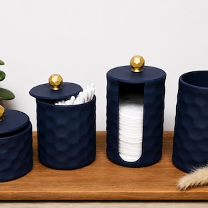 May include: A set of four navy blue bathroom storage containers with a textured honeycomb pattern and gold-colored spherical knobs on the lids. The set includes two lidded jars, one open-front cotton pad dispenser, and one open-top cylindrical holder.