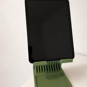 Puede incluir: Un soporte para tableta de color verde salvia con una base de múltiples ranuras para ángulos de visión ajustables y una pequeña bandeja rectangular en la parte frontal. Una tableta negra está colocada verticalmente con pantalla oscura.