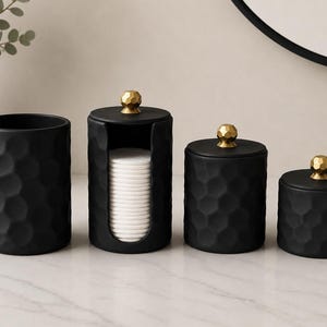May include: A set of four matte black cylindrical containers with a textured honeycomb pattern. Three containers feature removable lids with gold-toned, faceted spherical knobs. One container is an open-front dispenser holding white cotton pads.