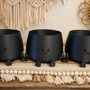May include: Three matte black planters, each featuring a simple embossed face with two circular eyes and a curved mouth. These round containers are supported by small, stubby legs extending from the base, giving them a seated appearance.