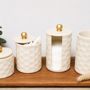 May include: A set of four cream-colored ceramic bathroom containers with a textured honeycomb pattern and gold-toned spherical knobs on the lids. The set includes two lidded jars, a cylindrical cotton pad dispenser with a vertical opening, and a cup.