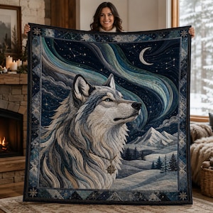 Op de afbeelding: Een rechthoekige quilt met een gedetailleerde illustratie van een grijze wolf met een kompas-hanger, tegen een besneeuwd berglandschap onder een maansikkel en wervelend blauw en groen noorderlicht, omlijst door een donkerblauwe sneeuwvlokrand.