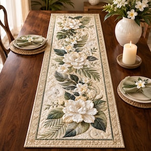 May include: A rectangular table runner featuring a quilted design with large white flowers and green tropical leaves on a cream background. The runner has a decorative border and is displayed on a wooden table with matching napkins and a candle.