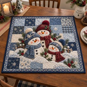 May include: A square fabric wall hanging or table mat featuring a quilted patchwork design in shades of blue and white. The center displays three plush snowman figures wearing knit hats and scarves, accented with pine branches and red berries.