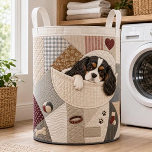 Könnte beinhalten: Ein zylindrischer Wäschekorb aus Stoff mit gestepptem Patchwork-Design in neutralen Beige-, Grau- und Kastanientönen. Die Außenseite zeigt applizierte Motive wie ein Herz, einen Knochen, ein Halsband und Pfotenabdrücke, mit einem Cavalier King Charles Spaniel.
