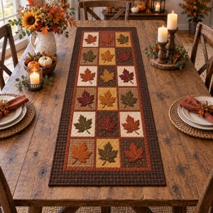 May include: A rectangular fabric table runner featuring a grid pattern of quilted maple leaf appliques in shades of orange, red, brown, and green. The runner has a dark brown plaid border and is centred on a wooden dining table with place settings.