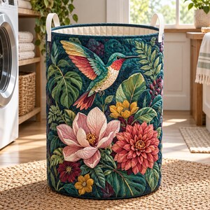 Op de afbeelding: Een cilindrische gewatteerde wasmand met een kleurrijke kolibrie in vlucht, omringd door grote roze magnolia's, rode dahlia's, gele bloemen en weelderig groen tropisch blad op een donkerblauwe achtergrond met twee witte handvatten.