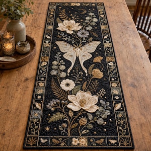 May include: A rectangular table runner featuring a black fabric base with intricate embroidery of a large central moth, white lotus-like flowers, ferns, and celestial motifs including stars and a crescent moon, finished with a decorative border.