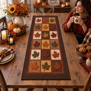 May include: A rectangular fabric table runner featuring a grid pattern of alternating squares. Each square displays an embroidered maple leaf in shades of orange, red, yellow, and brown, set against cream, brown, and patterned fabric backgrounds.