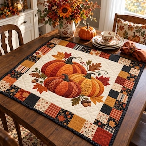 May include: A square quilted table topper featuring a central appliqué of three pumpkins surrounded by autumn leaves and acorns. The border consists of a patchwork of squares in shades of orange, brown, black, and cream with various autumn patterns.