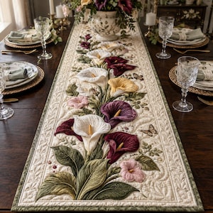 May include: A rectangular quilted table runner featuring an appliqued floral design of white, yellow, pink, and deep purple calla lilies with green leaves and small butterflies on a cream-colored background with intricate stitched detailing.