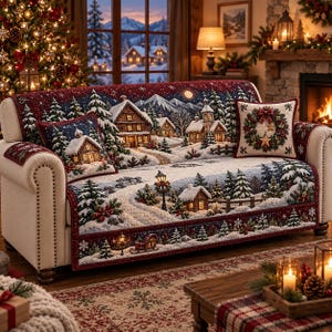 May include: A cream-colored sofa covered with a quilted throw featuring a winter village scene with snow-covered cabins, pine trees, and a red border. Two matching square throw pillows are included, one displaying a cabin and one a wreath.