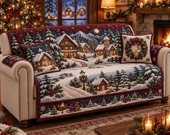 May include: A cream-colored sofa covered with a quilted throw featuring a winter village scene with snow-covered cabins, pine trees, and a red border. Two matching square throw pillows are included, one displaying a cabin and one a wreath.