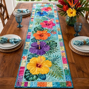 Op de afbeelding: Een rechthoekige gewatteerde tafelloper met een levendige tropische print van grote hibiscusbloemen in geel, paars, oranje, roze en blauw, geaccentueerd met groene monsterabladeren op een lichtblauwe achtergrond, omrand door een kleurrijke patchwork-bies.