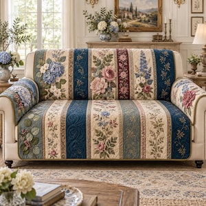 May include: A quilted sofa cover featuring a vertical striped pattern with floral motifs in shades of blue, cream, pink, and green. The fabric displays detailed botanical illustrations of hydrangeas, roses, and vines with a textured, stitched finish.