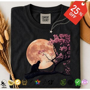 May include: A black crew neck t-shirt featuring a graphic print of a black cat sitting on a tree branch against a large, glowing orange full moon. The tree has pink cherry blossoms. A Comfort Colors brand label is visible at the inner neckline.