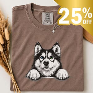 May include: A brown crew neck t-shirt featuring a graphic illustration of a black and white husky dog with one blue eye and one green eye. The dog is depicted with its front paws resting as if on an edge. The shirt has a Comfort Colors brand label.