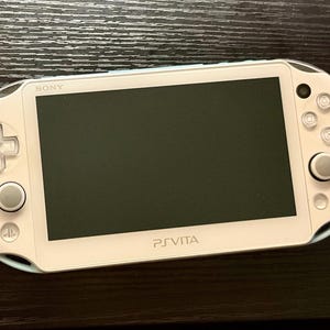 Könnte beinhalten: Eine weiße Sony PlayStation Vita Handheld-Spielkonsole mit einem rechteckigen schwarzen Bildschirm, einem Steuerkreuz, zwei Analogsticks, vier Aktionstasten mit Symbolen und dem PS Vita-Logo unter dem Bildschirm.