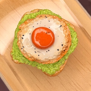 May include: A miniature food replica of a slice of toasted bread topped with a thick layer of bright green mashed avocado and a single fried egg. The egg features a glossy, vibrant orange yolk and a white with small black pepper specks.