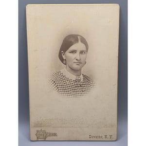 May include: A sepia-toned vintage cabinet card photograph featuring a portrait of a person with center-parted hair, wearing a checkered shirt with a white collar and a brooch. The bottom of the card displays the text "LUCAS" and "DUNKIRK, N. Y."
