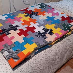May include: A handmade patchwork quilt featuring a repeating plus sign pattern. The quilt is constructed from various fabric squares in solid colors including red, orange, yellow, blue, and pink, alongside patterned fabrics with polka dots and gingham.