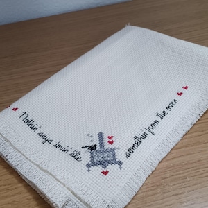 May include: A white rectangular kitchen towel with fringed edges featuring a cross-stitch design. The embroidery includes a grey stove icon, small red hearts, and the black text: Nothin' says lovin' like somethin' from the oven.