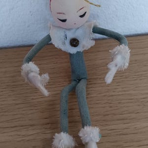 May include: A fabric elf ornament featuring a red pointed hat with a gold hanging string, a pale face with closed eyes, and yellow hair. The figure wears a grey body suit, a white ruffled collar with a brown button, and white fuzzy cuffs and boots.