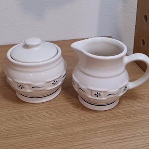 May include: A white ceramic sugar bowl with a lid and a matching creamer pitcher. Both pieces feature a rounded shape with a textured basket-weave pattern around the center, accented by small blue floral motifs and thin blue horizontal lines.