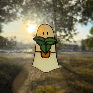 May include: A stained glass suncatcher shaped like a ghost with a pale off-white body and two small black eyes. The ghost is holding a small orange potted plant with green leaves, constructed with dark metal solder lines and hanging by a thin wire.