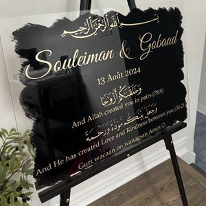 Peut inclure: Un panneau de mariage rectangulaire en acrylique transparent avec un centre peint en noir et une calligraphie dorée. Il affiche les noms Souleiman & Gobaad, la date du 13 Août 2024, des versets en arabe et en anglais, et Guri wacaan oo wanagsan Amin.