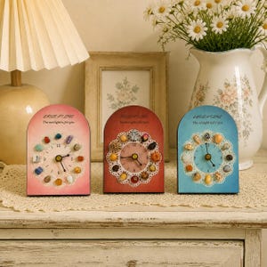 Puede incluir: Tres relojes de mesa arqueados con el texto "SMILE AT LIFE The sunlight is for you" impreso en la parte superior. El reloj de la izquierda es rosa con piedras coloridas, el central es rojo con piñas pequeñas y el de la derecha es azul con conchas.