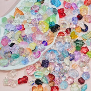 Puede incluir: Una gran colección de cuentas acrílicas translúcidas variadas en diversas formas, incluyendo flores, estrellas, corazones y figuras geométricas. Las cuentas presentan un espectro de colores vibrantes como rosa, azul, amarillo, verde y morado, con algunas piezas transparentes.