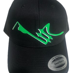 Puede incluir: Una gorra snapback negra con un gráfico estilizado de color verde brillante en el panel frontal. La gorra tiene una corona estructurada, una visera plana y una parte trasera de malla. Una pegatina circular en la visera dice The Authentic Yupoong Snapback Yupoong Since 1974.