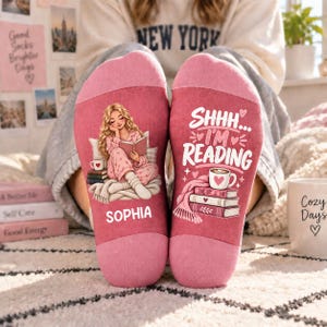 May include: A pair of pink socks featuring a graphic of a person reading a book on the left and the text SHHH... I'M READING above a stack of books and a coffee mug on the right. The name SOPHIA is printed in white on the left sock.