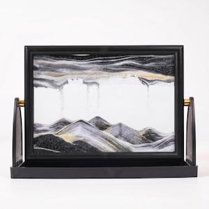 May include: A rectangular moving sand art display featuring a black frame with gold-toned side pivots. The glass panel contains layers of black, white, and gold sand forming mountain-like peaks at the bottom and cloud-like formations above.