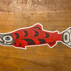 Op de afbeelding: Een sticker met een gestileerde illustratie van een zalm in levendige rode en zwarte tinten. Het ontwerp bevat traditionele formline-kunstelementen met een witte omlijning, met een profiel met scherpe tanden en een staartvin.