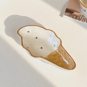 May include: A ceramic dish shaped like an ice cream cone with a cream-coloured top featuring small brown dots and a textured brown waffle cone base. The dish has a glossy finish and a brown rim that outlines the entire ice cream cone silhouette.
