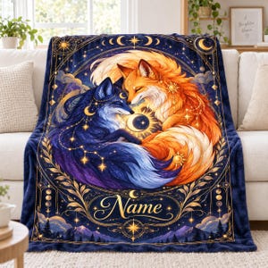 May include: A plush throw blanket featuring a dark blue background with a large circular design of a blue fox and an orange fox curled together. The design includes gold celestial motifs, stars, moons, and the word Name in a cursive script.